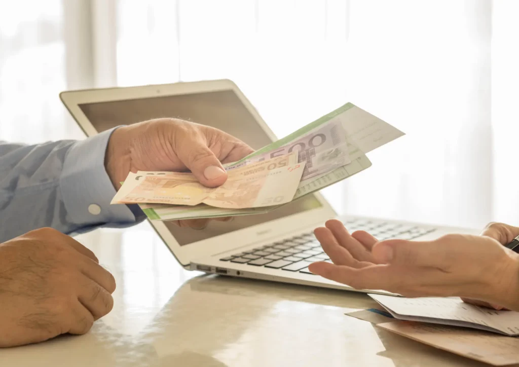 Close-up of hands exchanging money over a laptop, symbolizing the cost and investment required for lead generation.
