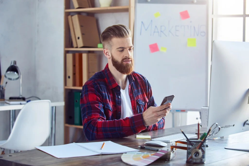 "Digital marketer analyzing cost per lead metrics on his phone at his desk in a creative marketing office."